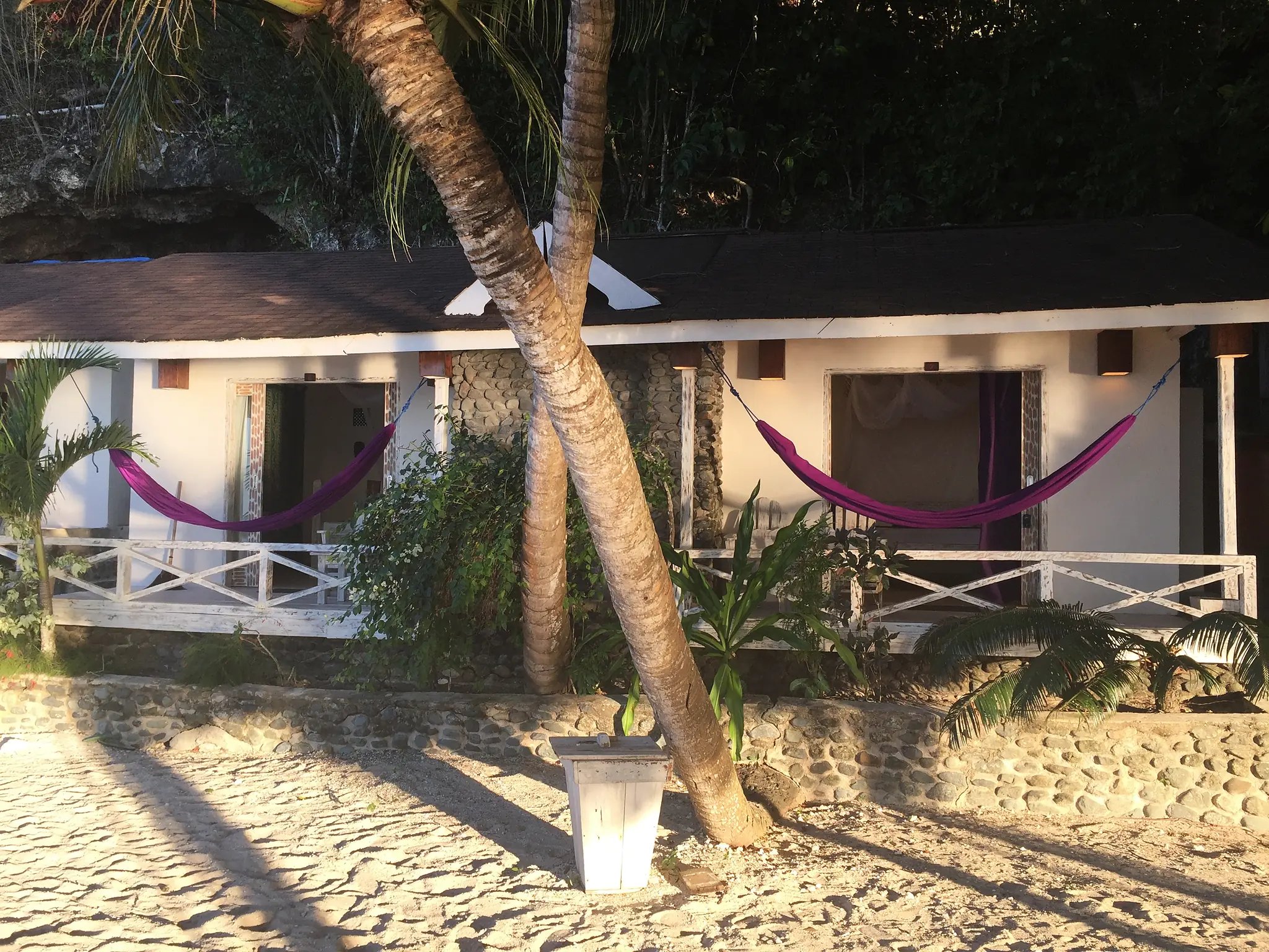 the row of deluxe beachfront bungalows on the sand under palms at dusk, hammocks on each veranda